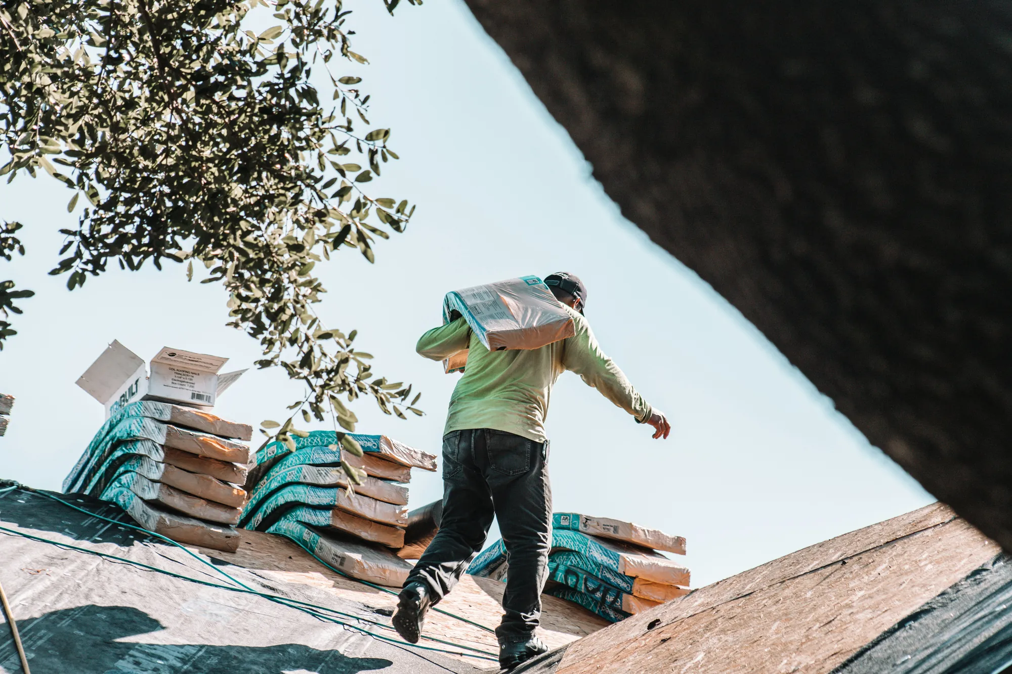 A roofer carrying a bundle of shingles across a roof being replaced