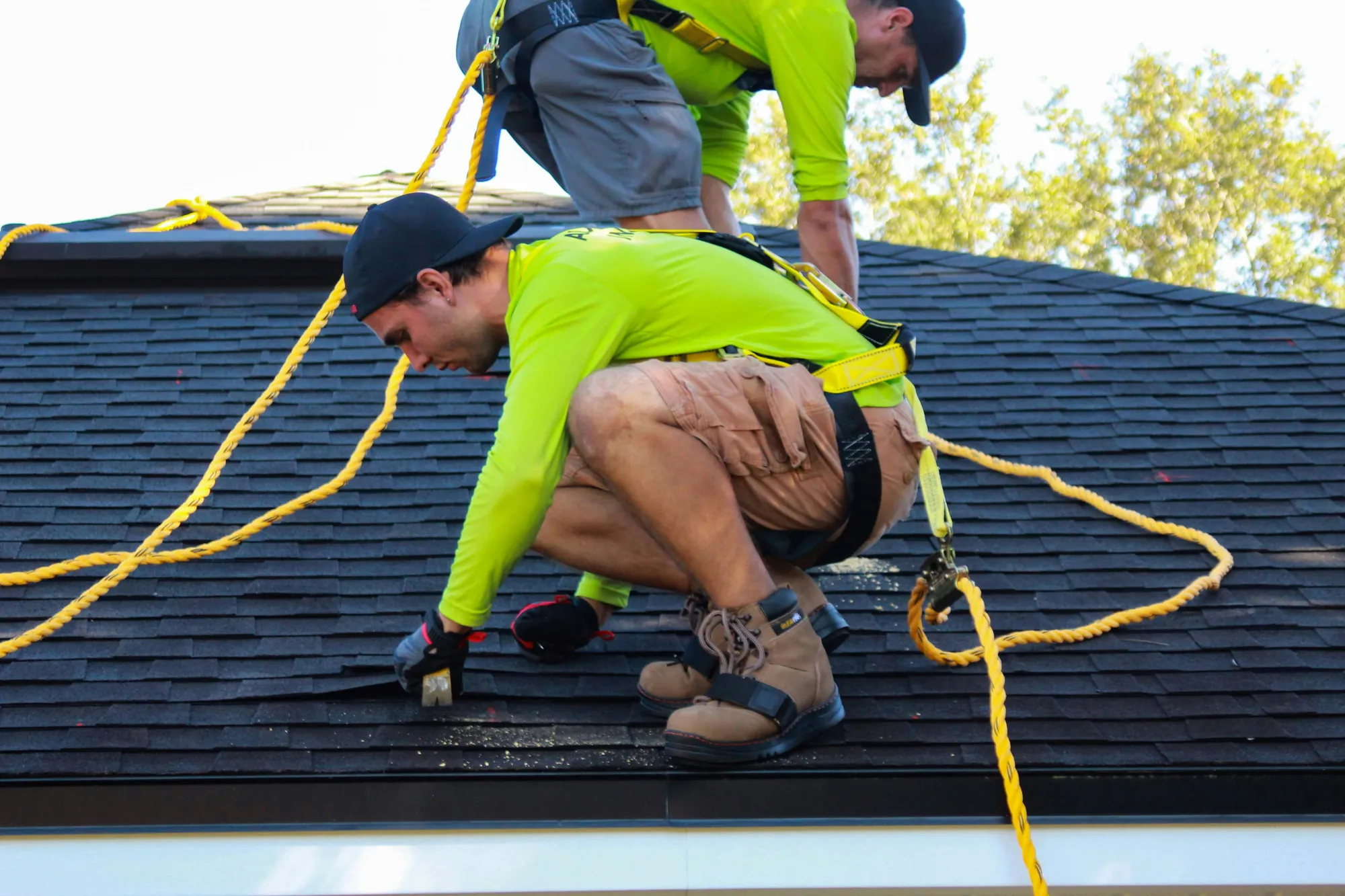 A roofer lifting a damaged shingle with a pry bar while a second roofer works behind him