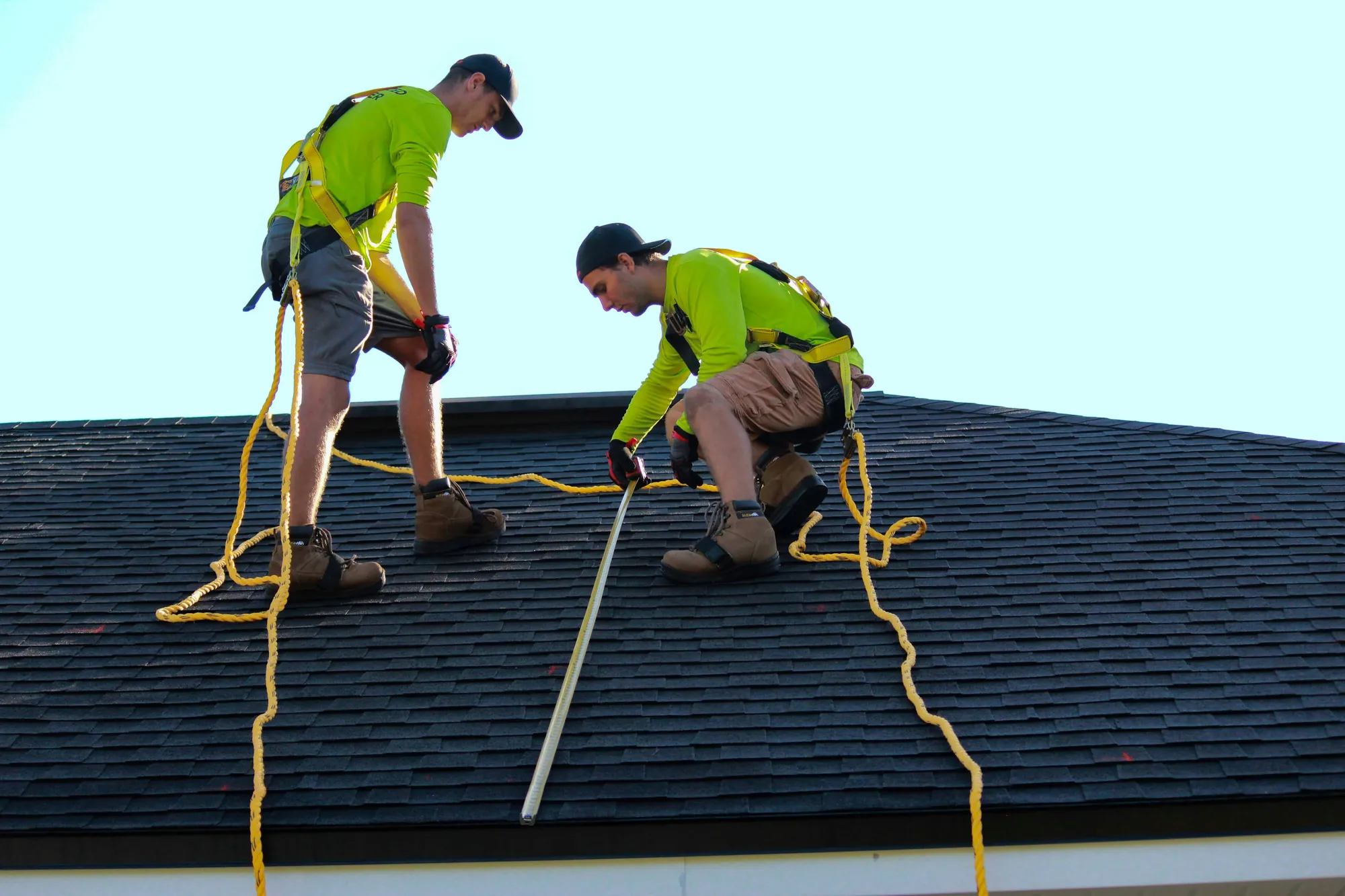 Two roofers in safety harnesses measuring a shingle roof